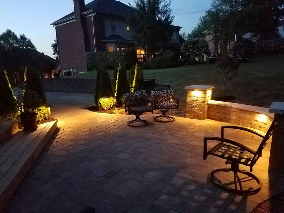 A paver patio at dusk with a stacked-stone pillar and built-in lighting, landscape lighting along a retaining wall and shrubs, outdoor furniture arranged for entertaining.